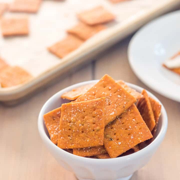 small white bowl filled with homemade gluten free pizza crackers on a wooden table with a pan of gluten free crackers in the background