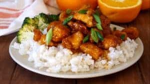 white plate with rice, orange chicken, green onions and broccoli with an orange in the background
