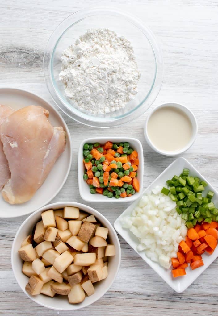 raw chicken, diced potatoes, choped carrots, chopped onion, chopped celery, frozen peas and carrots, egg and milk, all in small bows and dishes on a white background