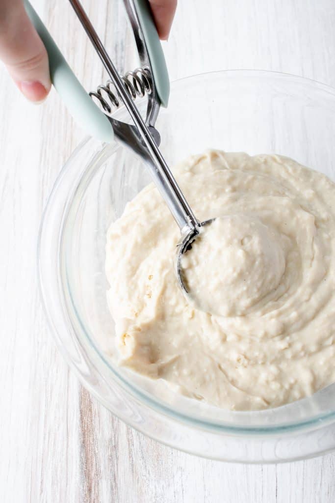 silver spring loaded scoop scooping gluten free dumpling batter in a glass bowl on a white wooden backdrop