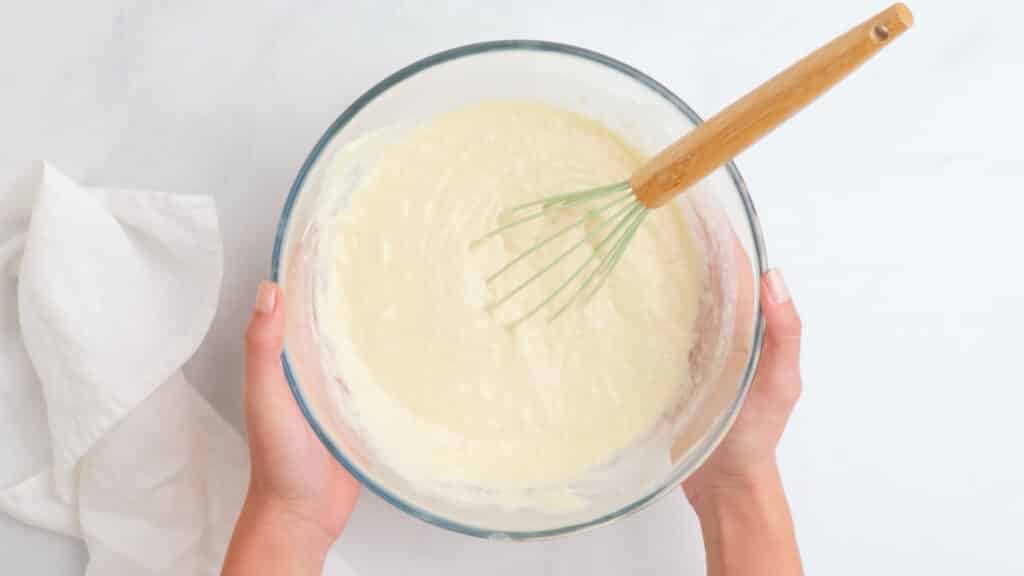 glass bowl with gluten free waffle batter and a whisk with a wooden handle on a white table