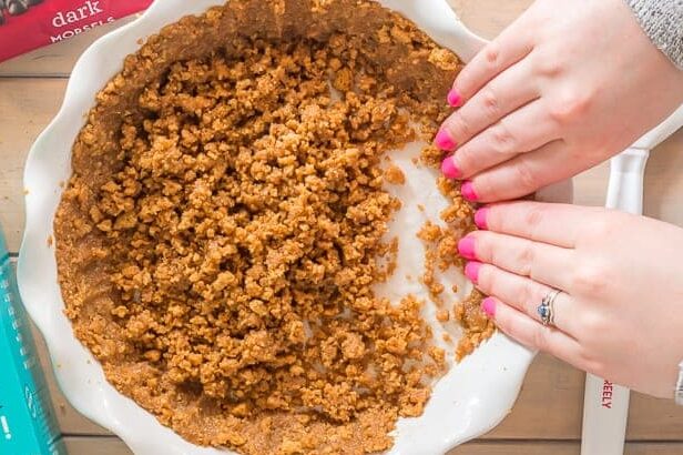 hands with pink painted finger nails pressing a gluten free graham cracker crust up the sides of a white ceramic pie plate with a box of cookies and bag of dark morsels on the wooden table