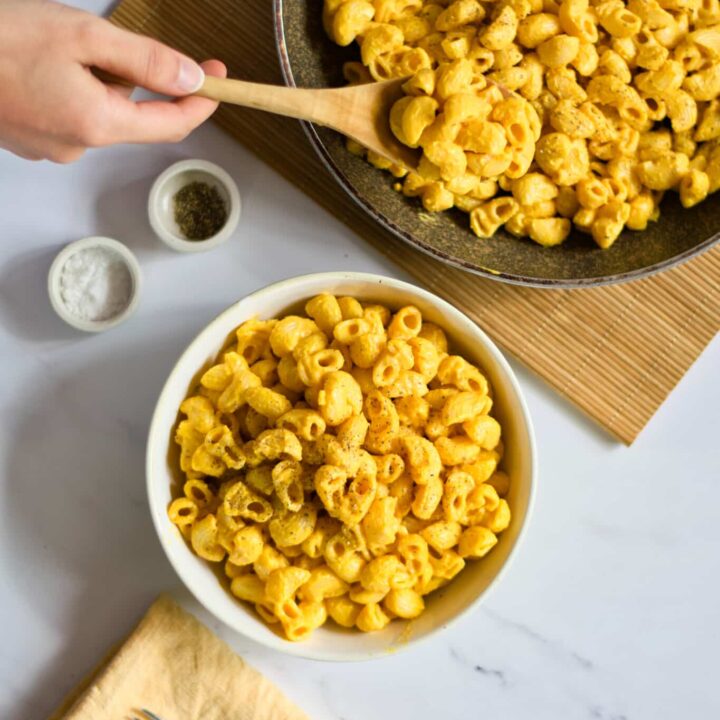 overhead view of vegan macaroni and cheese with dairy free cheese sauce and gluten free pasta being served from a brown bowl with a wooden spoon into a white bowl on a marble countertop