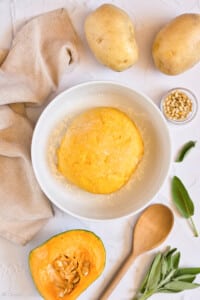 a bowl filled with gluten free pumpkin gnocchi dough on a white table with butternut squash, potatos, sage, and pine nuts on it