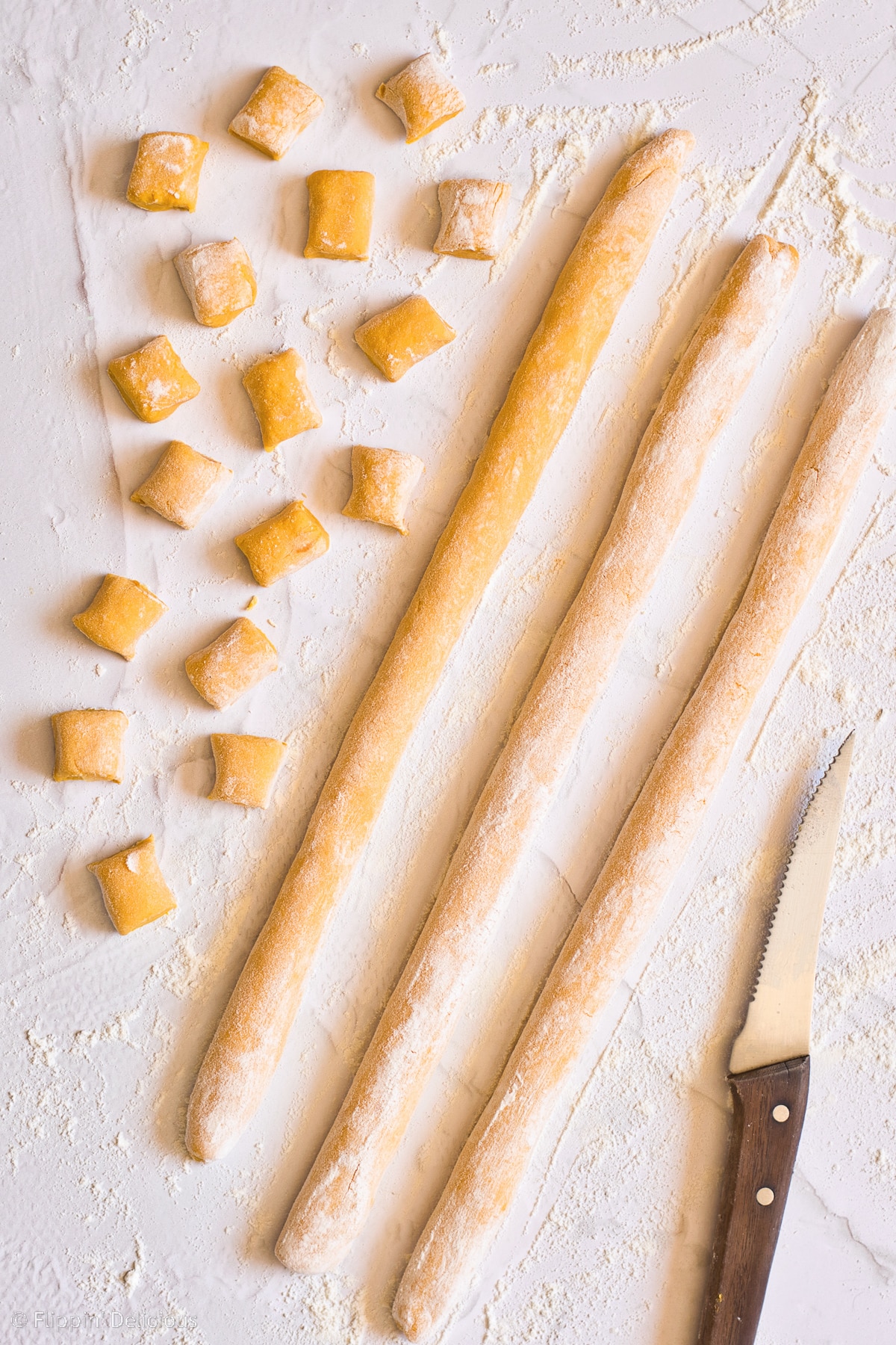 Cut pieces of gluten free gnocchi dough on a white marble table ready to be shaped with a knife beside them with a wooden handle