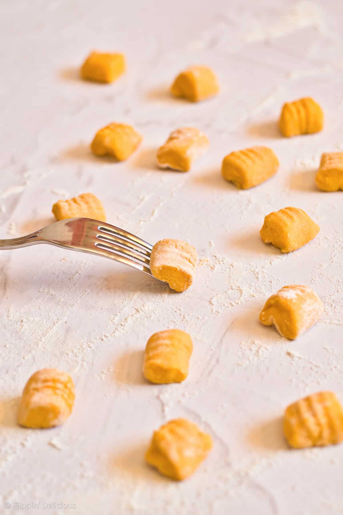 rolling gluten free gnocchi over the upside down tines of a fork, on a white marble floured countertop with a bunch of shaped vegan pumpkin gnocchi already shaped beside it.