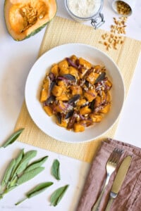 bowl of vegan pumpkin gnocchi on a wooden place mat with a brown napkin on the table with silver cutleryu, sage leaves, pine nuts, and butternut squash on the table