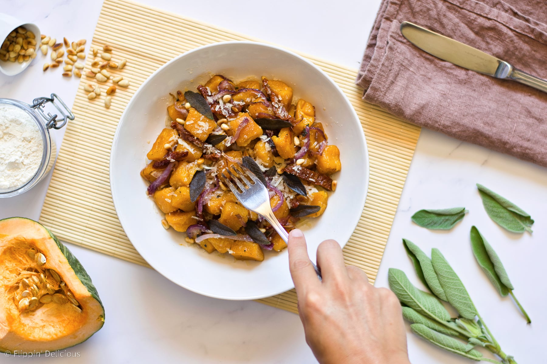 overhead photo of a fork reaching into a white bowl with gluten free pumpkin gnocchi with pine nuts sundried tomatoes and sage on a wooden rolled placemat