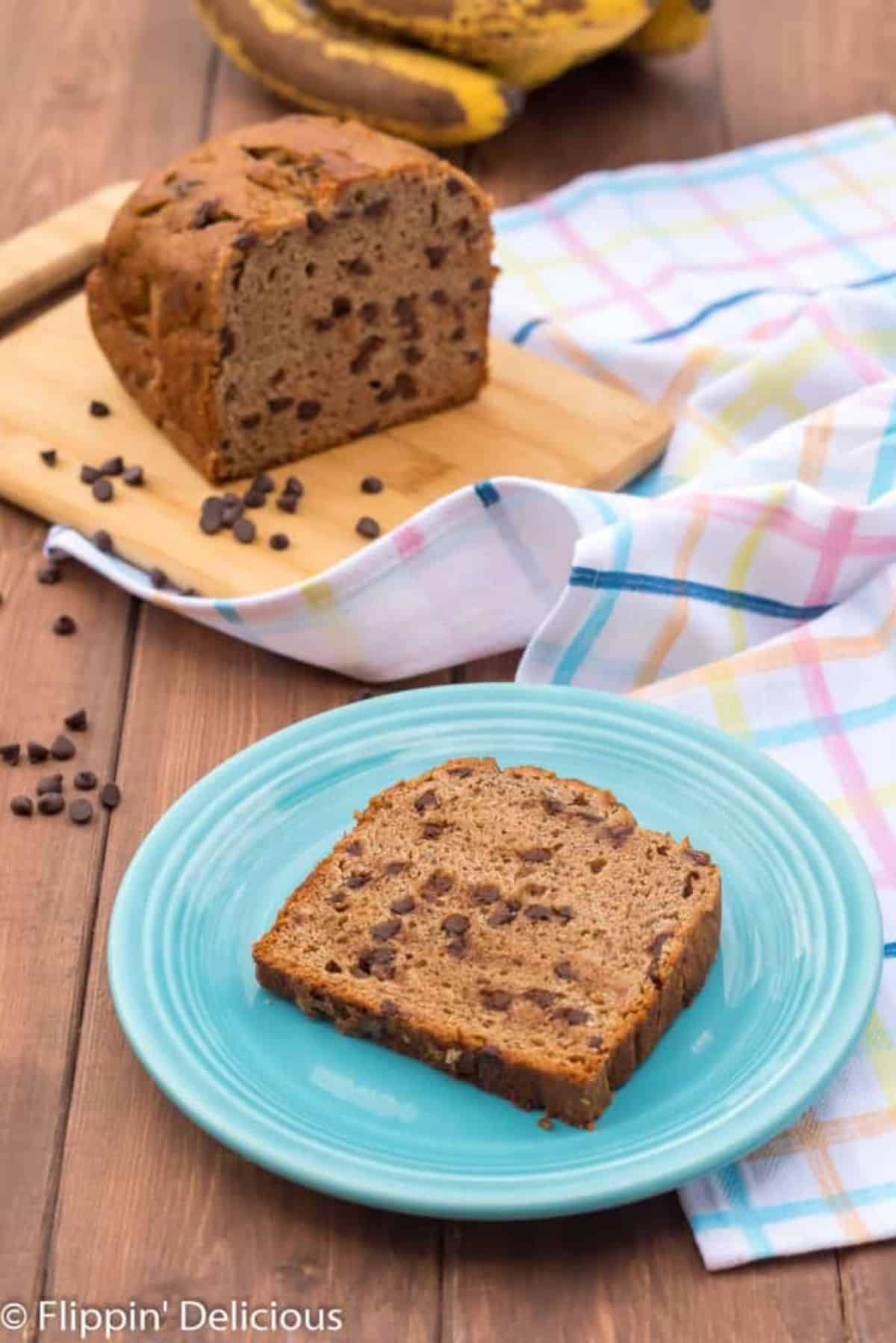 A loaf of Gluten-free Chocolate Chip Banana Bread on a wooden cutting board, a slice of Gluten-free Chocolate Chip Banana Bread on a blue  plate.