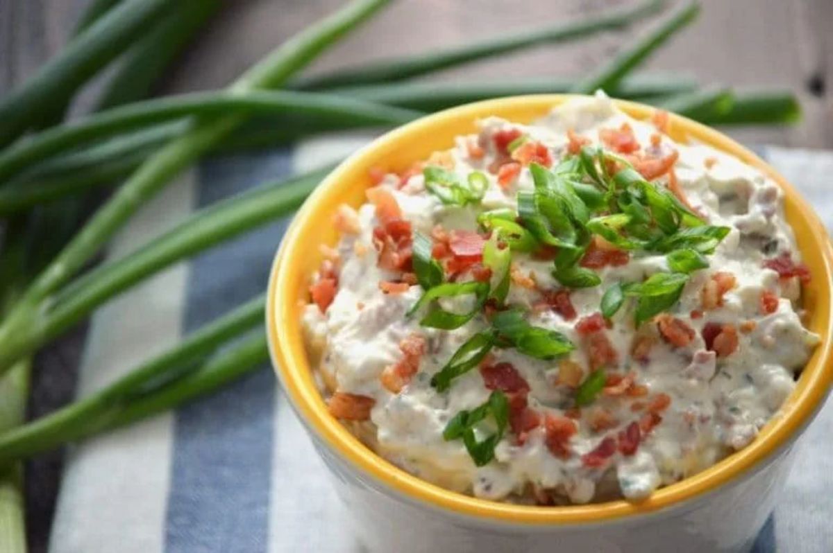 Crack Dip in a white-yellow bowl with bacon and herbs on the top.