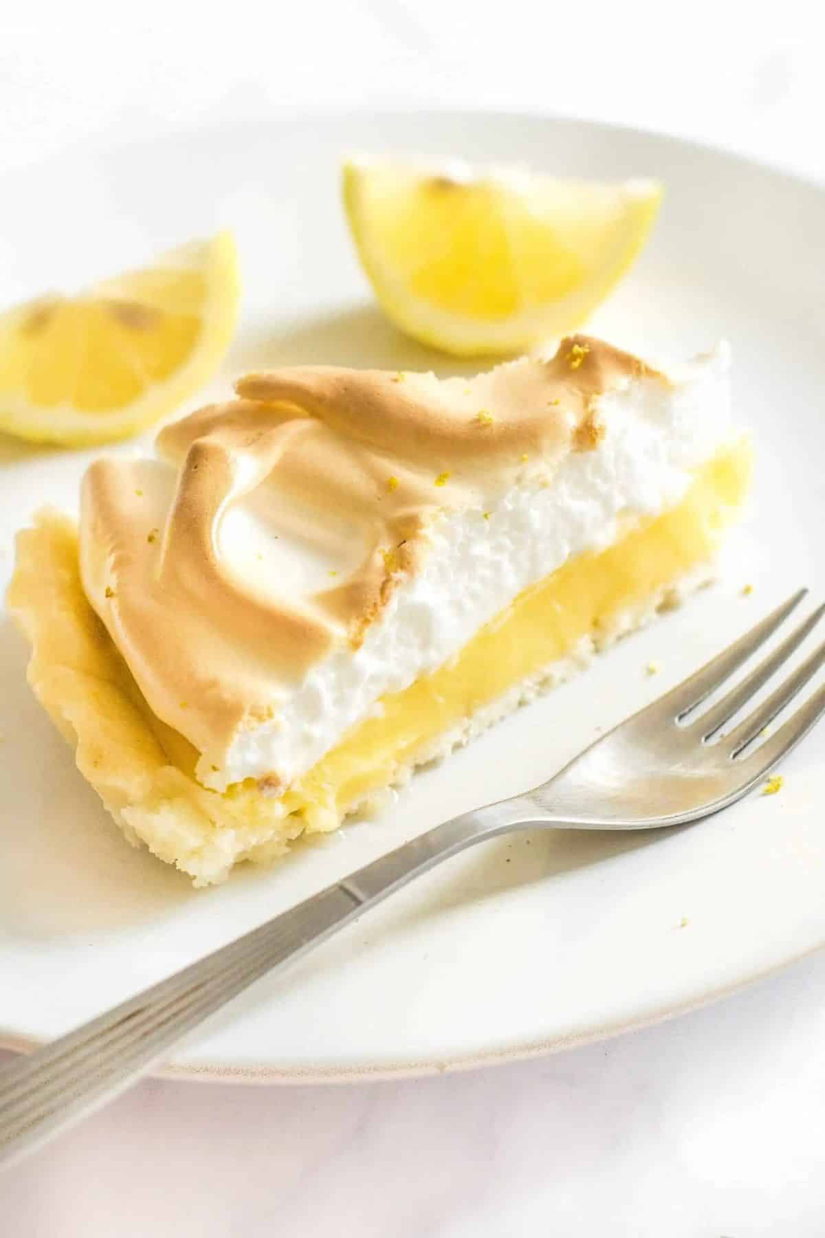 A piece of Lemon Meringue Pie on a white plate with a fork and two piece of lemon.