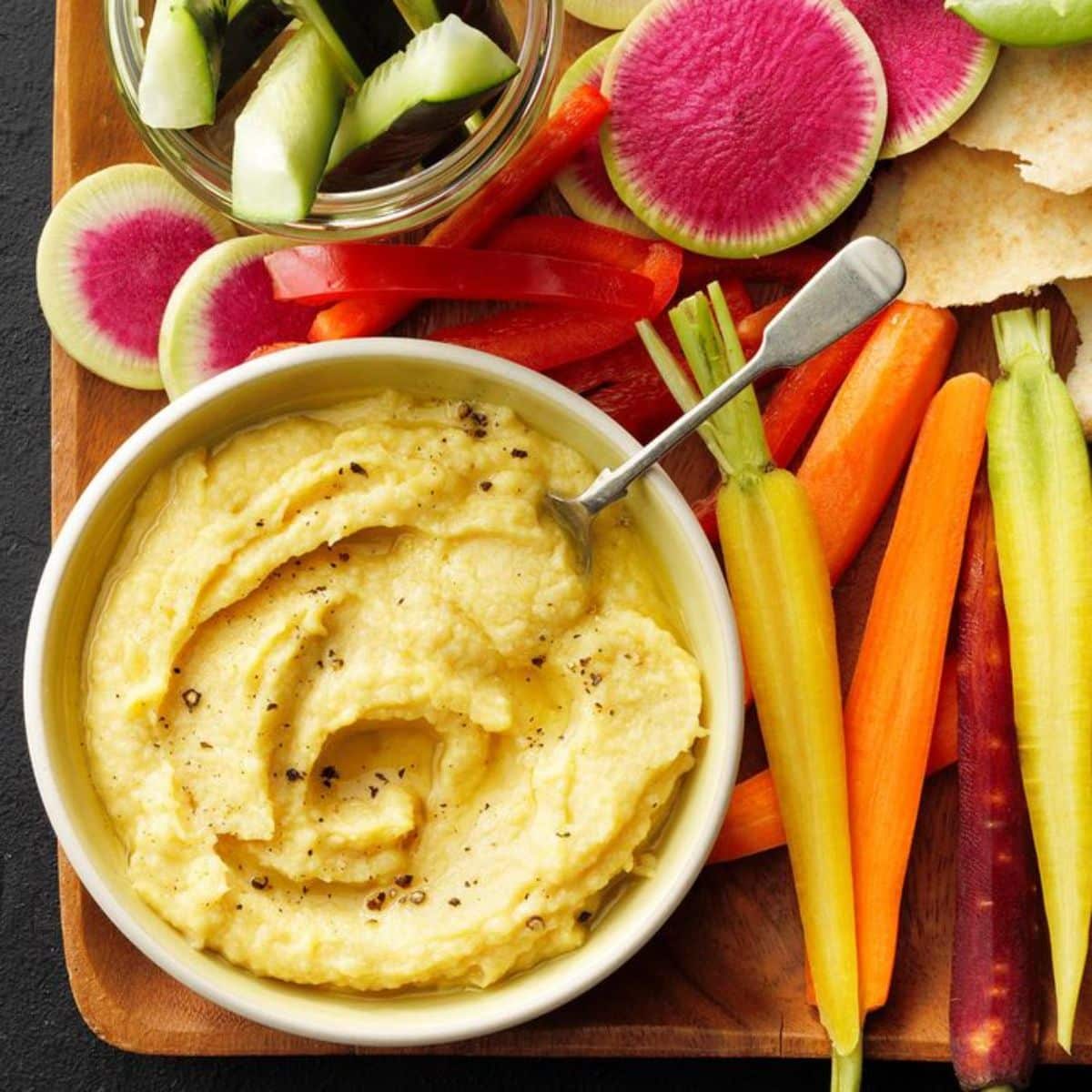 Lemon Garlic Hummus in a white bowl with a spoon on a wooden tray with veggies.