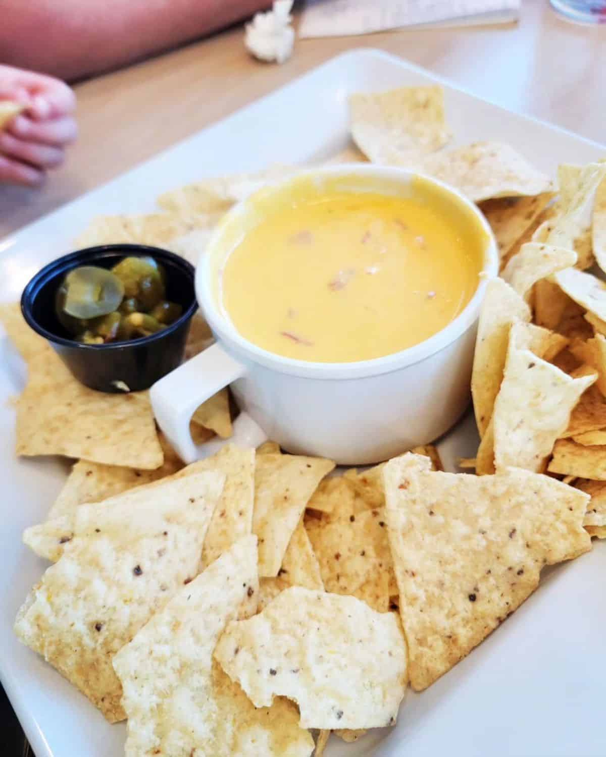 Gluten-Free Nacho Cheese Dip in a white cup on a tray with nachos.,
