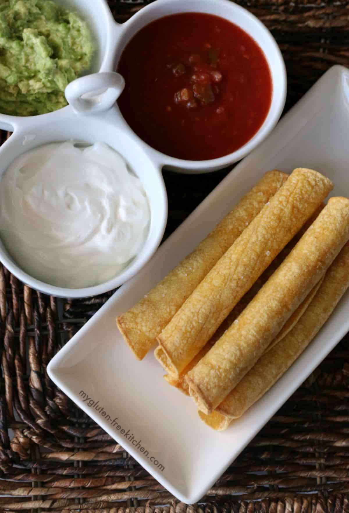 Delicious gluten-free Baked Taquitos on a white tray next to bowls full of different dips.