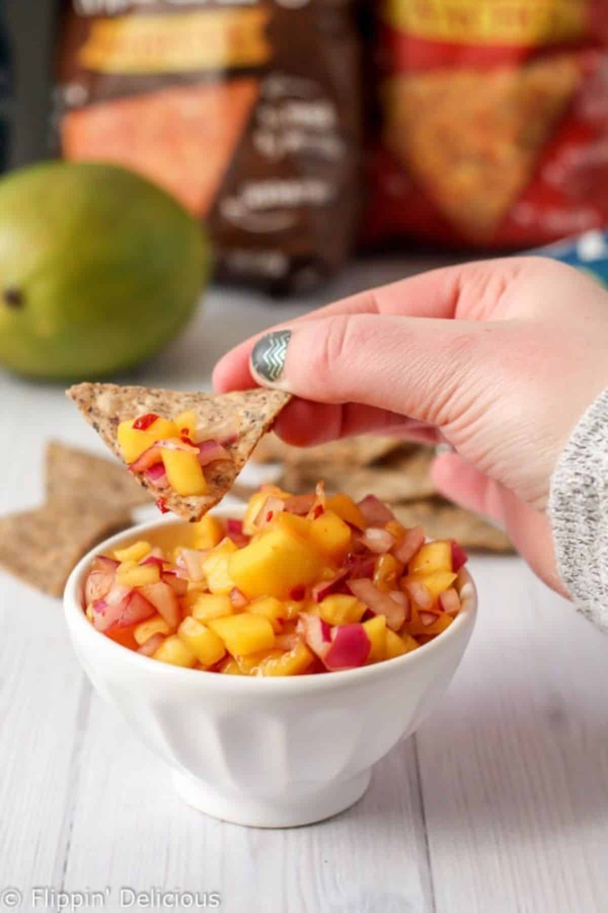A cracker dipped in Chipotle Mango Salsa over a bowl of Chipotle Mango Salsa.