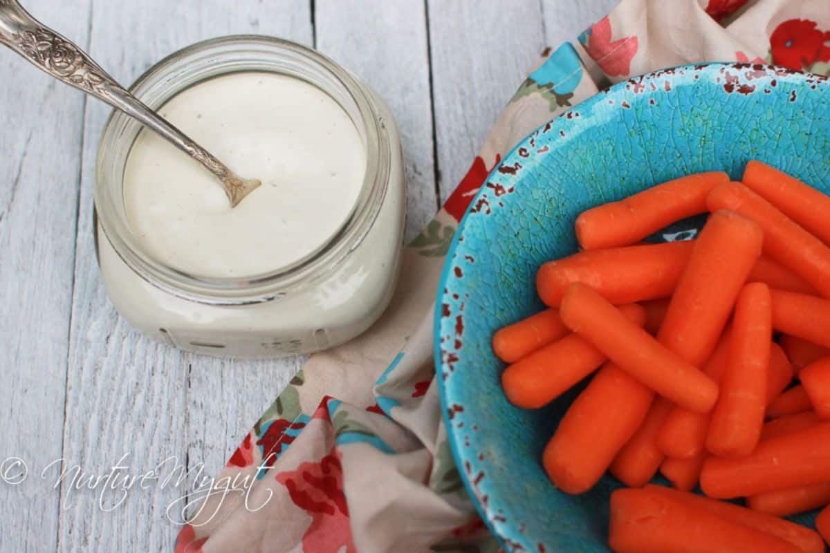 Tasty yDairy-Free Sour Cream in a glass jar with a spoon next to a bowl of baby carrots.